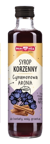 Szklana przezroczysta butelka z czarnym zakręcanym korkiem, wypełniona syropem w kolorze ciemnofioletowym z etykietą „Syrop korzenny Cynamonowa aronia” i ilustracją aronii i cynamonu. Przeznaczony do herbaty, wody, grzańca.